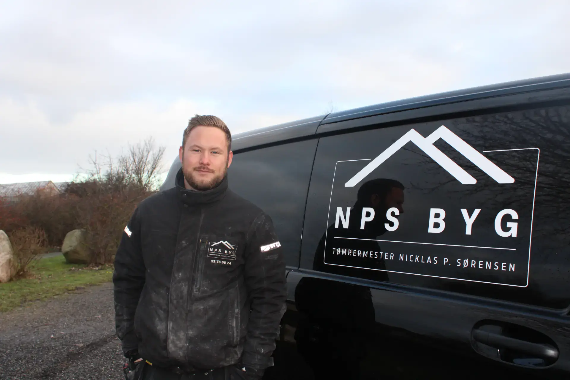 Mand står foran NPS Byg firmabil med logo, udendørs setting.