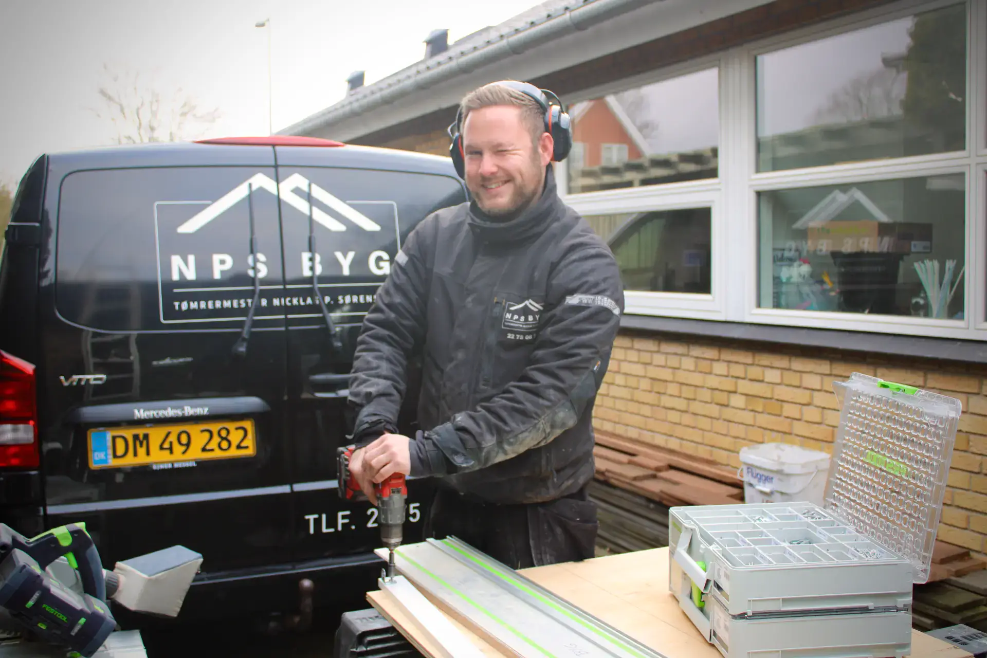 Billedet viser en smilende tømrer, der arbejder udendørs med værktøj og udstyr foran en NPS Byg vare.