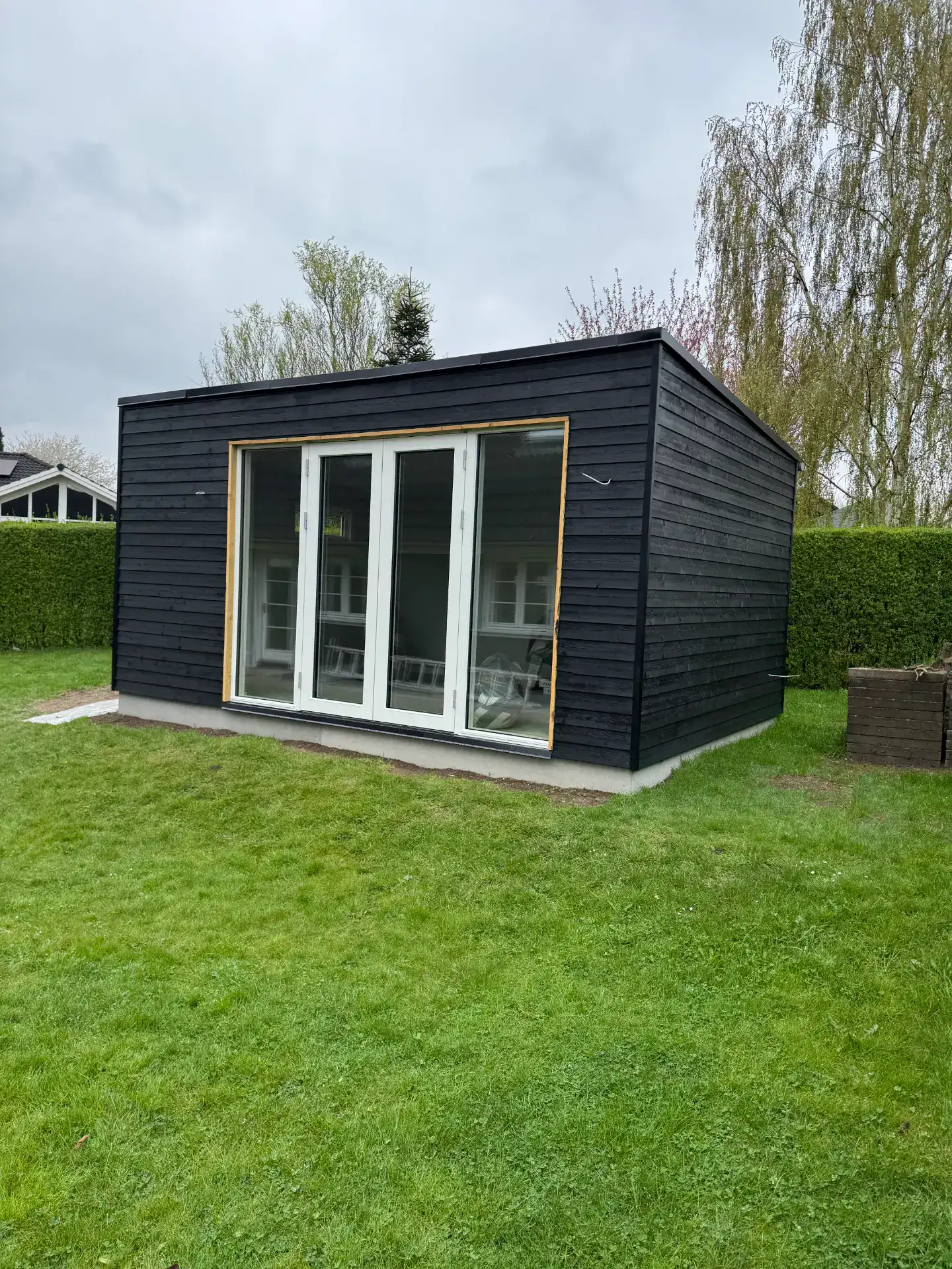 Moderne sort træhytte med store glasdøre i en grøn have.